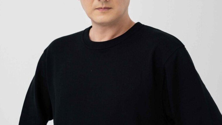 Retrato de divulgação do ator e dublador japonês Hiroshi Tsuchida, de camiseta preta diante de um fundo claro