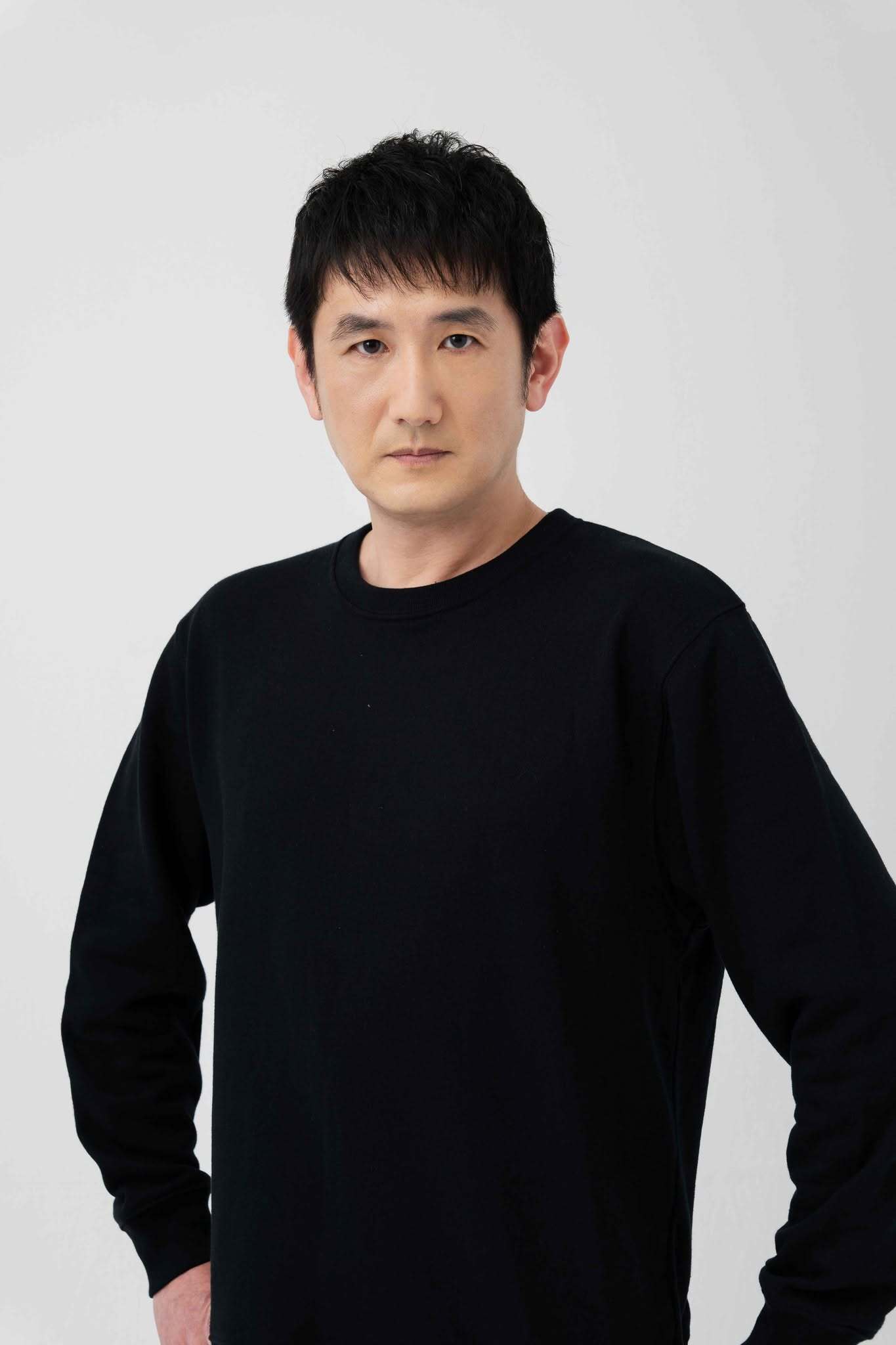Retrato de divulgação do ator e dublador japonês Hiroshi Tsuchida, de camiseta preta diante de um fundo claro