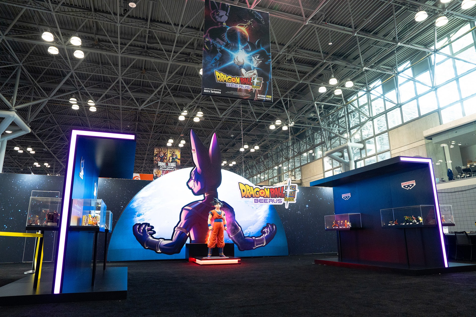 Estande de Dragon Ball Super: Beerus na NYCC 2026, com painel de Beerus, estátua de Goku e vitrines de figuras