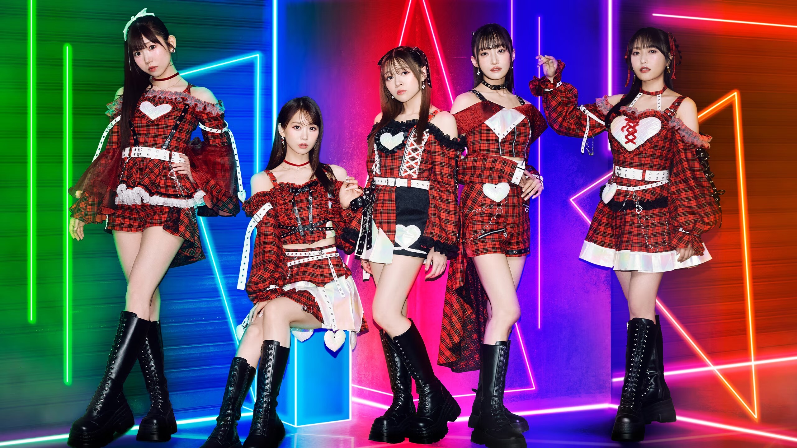 As cinco integrantes do i☆Ris com figurinos xadrez vermelhos e pretos diante de luzes neon coloridas
